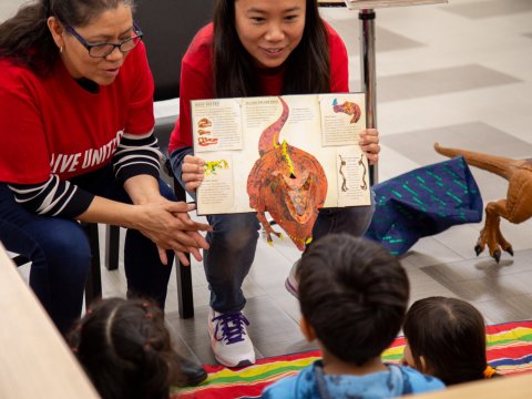 Two women show off a pop up dinosaur book to two kids